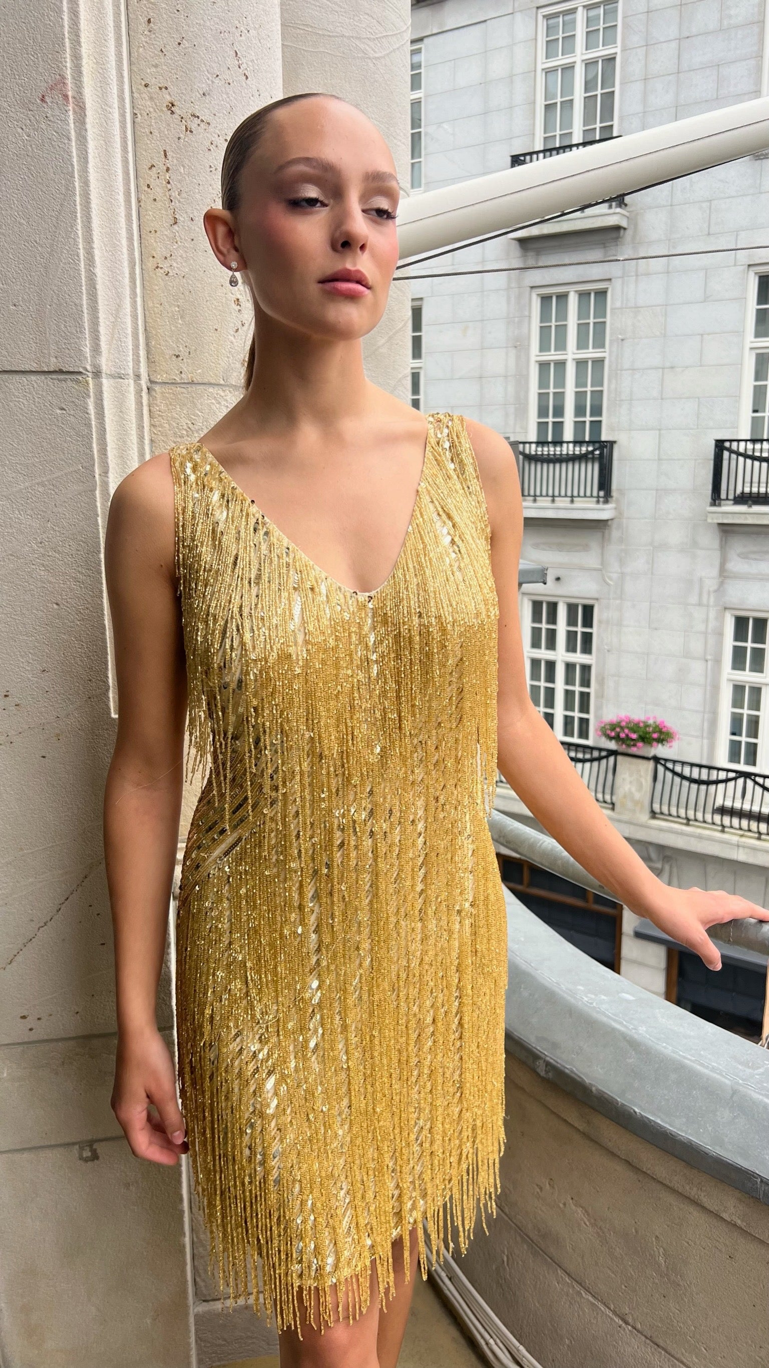 Woman wearing a gold fringe dress standing against a building exterior.